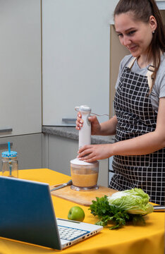 Adult Girl Holds Blender While Mixing The Ingredients For Smoothie And Looks At Laptop That Is On The Table. Healthy Eating. Selective Focus.