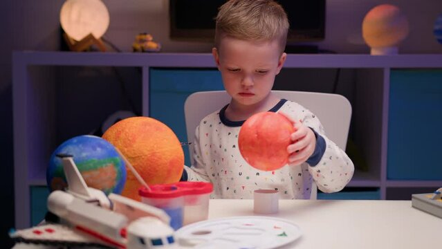 Blond boy paint planet solar system Mercury with colorful paint sitting home table in evening, planet solar system, spaceships and space shuttle from constructor around. Cosmonautics Day on April 12.