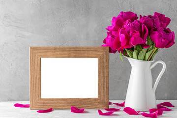 Blank photo frame and pink peony flowers with water drops in a vase on white wooden table. Still life
