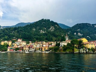 Naklejka na ściany i meble Varenna view, Italy. Colourful coastal houses view from Como lake. Hills and mountains. Forest, park, cloudy day, yellow buildings, bell tower.