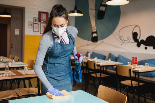 young waitress with apron, face mask and gloves cleaning tables restaurant with sanitizer and cloth