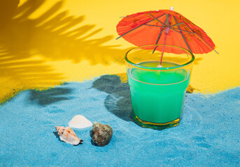 cocktail glass with green drink and red parasol on blue beach sand with sea shall under the tropical palm leafs on yellow background. creative minimal composition. © Nenad