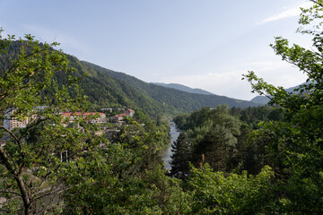 mountain landscape with a river with blue water, red roofs on the left and green trees and shrubs in the foreground, beautiful mountain landscape with blue skies in the distance