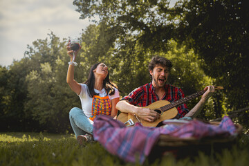 young people singing guitar at picnic - couple drinking wine in the countryside - millennials carefree have fun together on the summer weekend outing