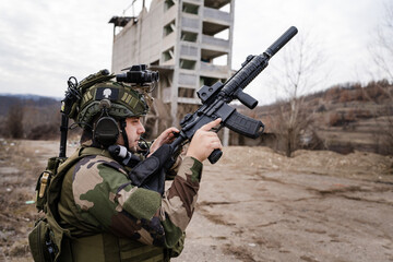 One man soldier armed with rifle wearing camouflage uniform loading gun while standing in front of ruined building in battle zone dogs of war professional mercenary or volunteer side view copy space