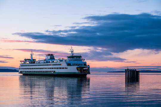 A large boat travels across the water at sunset with a colorful sky behind and clouds above