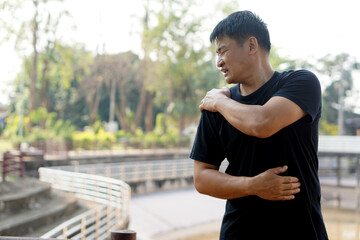 Asian man hurts his shoulder, stands at outdoor park. Concept : Health problems, swelling and...