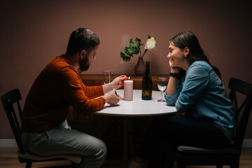 Hansome bearded young man lighting candle with match during talking with girlfriend while sitting together at table. Love couple celebrating anniversary or Valentines day having romantic dinner.