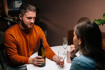 High-angle view of young man pouring wine into glass during talking with wife sitting together at festive table. Love couple celebrating anniversary or Valentine day having romantic dinner
