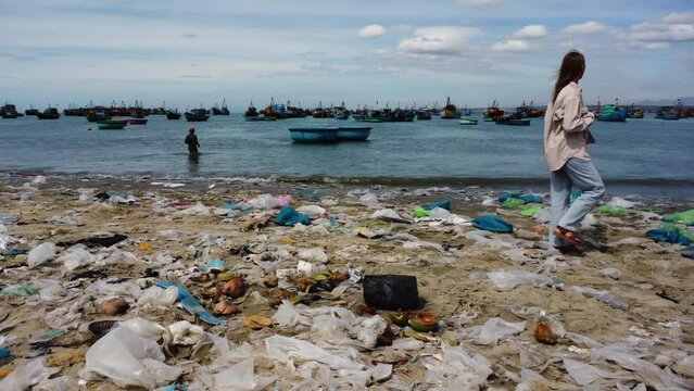 Sad young woman walking on sandy beach covered in piles of trash, pan right view