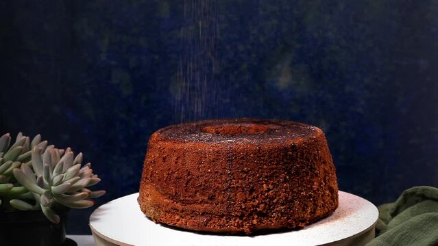 Chocolate cake being sprinkled with chocolate powder. Front image, green fabric and vase with succulent. Dark background.