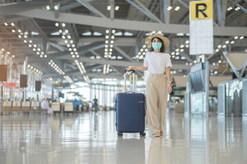 Young female wearing face mask with luggage walking in airport, protection Coronavirus disease infection, Asian woman traveler with hat. Time to travel after vaccine booster dose concept
