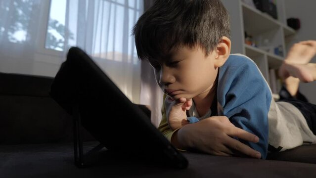 Asian boy watching computer tablet with addict expression on the couch. A problem for the kid who keeps using the screen.