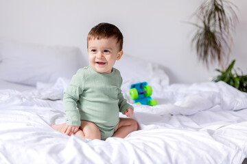 baby in a green cotton bodysuit is sitting on a white bed with a colored toy car. child is playing