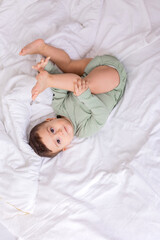 cute smiling baby in a green cotton bodysuit is lying on a white bed on his back. top view