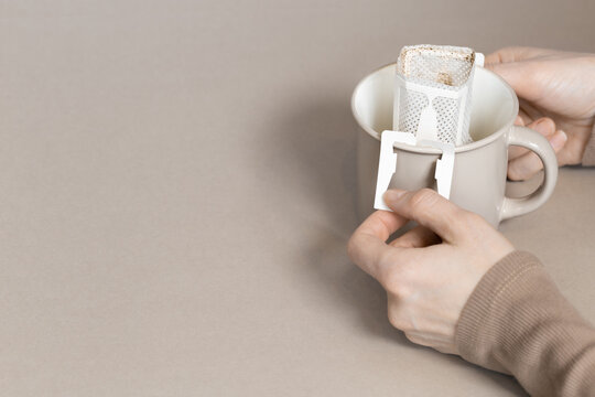Drip coffee bag with ground coffee in big cup, woman hands holding package with paper bag drip coffee filter for brewing on beige color background with copy space, monochrome colored.