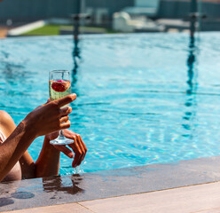 Female model is holding glass of wine in the pool with lovely seascape view, sunset