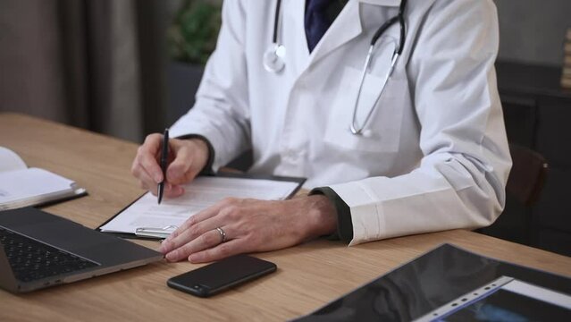 Closeup of male doctor writing prescription and sitting in front of laptop at table in clinic spbas. Middle aged man writes on paper how to treat disease and does diagnostic job, sits at desk with