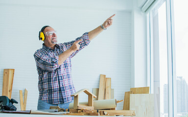 Senior Caucasian retired old male woodworker, carpenter pointing out window, wearing check shirt, safety headphone, standing with creating DIY wooden decoration, furniture as hobby after retirement.