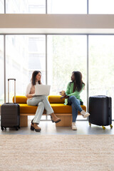 Young women waiting in hotel reception with trolley suitcases, talking, working with laptop. Vertical image.