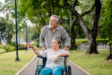 Fototapeta na wymiar Asian senior man with wife in wheelchair walking relaxing happy outdoors in the park, Leisure activities of retirees, elderly health care concept.