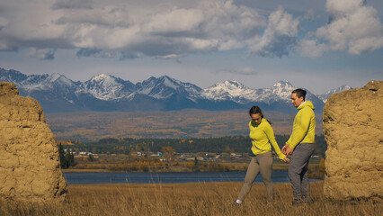 Man and woman in yellow green sportswear. Lovely couple of travelers hug and kiss near old stone enjoying highland landscape. Two travelers are walking against the backdrop of snow-capped mountains.
