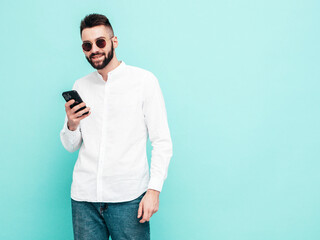 Handsome confident model.Sexy stylish man dressed in shirt and jeans. Fashion hipster male posing near blue wall in studio. Holding smartphone. Looking at cellphone screen. Using apps. Isolated