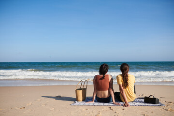 Fototapeta premium Young couple sitting on blanket on beach and looking at sea wave, view from the back