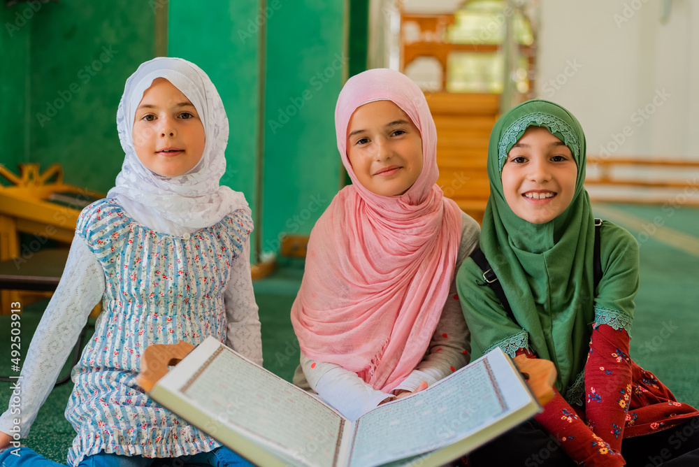 Wall mural Group of a children reading a holy book Quran in the mosque. Happy Muslim family. Muslim girls in hijab studying Islam religion.	