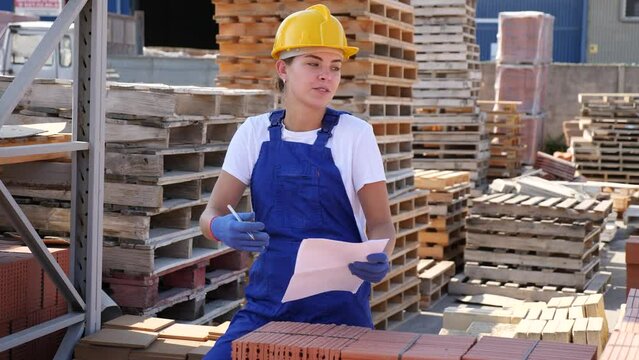 Young female worker Young woman working in construction material storage. She's writing something on sheet of paper while standing beside brick stacks in warehouse.writing beside brick stacks. High