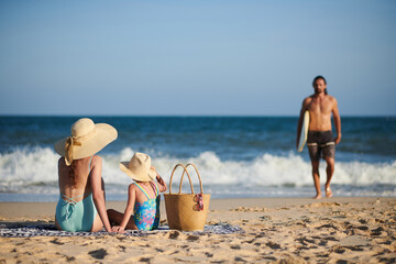 Woman in big straw hat and her daughter looking at husband returning after surfning in sea