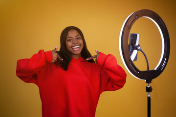 blogging, technology, emotions and people concept - happy african-american girl blogger in a red hoodie with a ring lamp and phone pointing fingers at herself on a yellow background with copy space