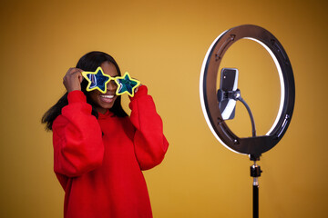 happy African-American girl blogger in a red hoodie and big star glasses with a lighted ring lamp and phone on a yellow background with copy space