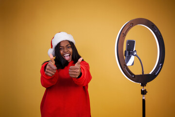 blogging, new year, technology and people concept - smiling african-american girl vlogger in a red hoodie and a Santa hat with a lighted ring lamp and phone showing thumbs up on a yellow background