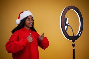blogging, new year, technology and people concept - smiling african-american girl vlogger in a red hoodie and a Santa hat with a lighted ring lamp and phone showing thumbs up on a yellow background