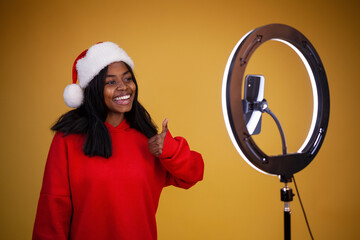 smiling african-american girl vlogger in a red hoodie and a Santa hat with a lighted ring lamp and phone showing thumbs up on a yellow background with copy space