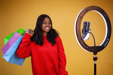 happy African-American girl in a red hoodie with colorful bags on her shoulder, a ring lamp and a phone on a yellow background with copy space