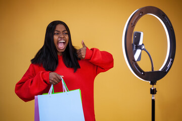 blogging, technology, advertisement, shopping and people concept - a joyful African-American vlogger girl with multicolored bags, a ring light and a phone showing thumbs up on a yellow background