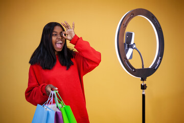 blogging, technology, advertisement, shopping and people concept - happy african-american girl vlogger in hoodie with colorful bags, ring light and phone showing ok sign on eye on a yellow background