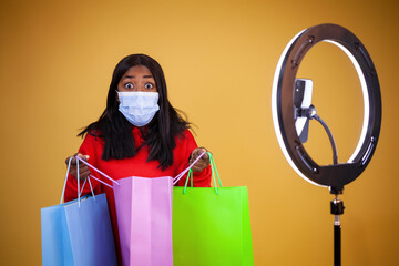 blogging, technology, pandemic, shopping and people concept - a surprised African-American vlogger wearing a protection mask opening colorful bags, with a ring lamp and phone on a yellow background