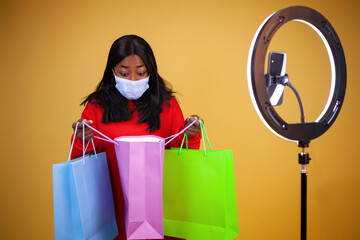a surprised African-American vlogger girl wearing a virus protection mask opening colorful bags, with a ring lamp and phone on a yellow background with copy space