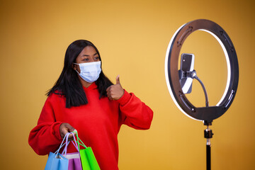 blogging, technology, pandemic, shopping and people concept - A happy African-American girl vlogger wearing a virus protection mask with colorful bags, a ring lamp and a phone showing thumbs up