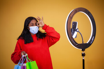 happy african-american girl blogger in virus protection mask with multicolored bags, ring lamp and phone showing ok sign on a yellow background with copy space