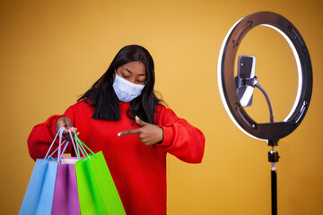 blogging, technology, pandemic, shopping and people concept - young african-american girl vlogger in a virus protection mask with a ring lamp and phone pointing finger at bags on a yellow background