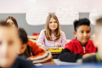 A smiling girl sitting at the desk in classroom with classmates.