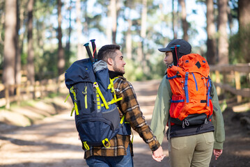 Side view of happy couple of young hikers. Caucasian man with beard and woman in cap with big backpacks holding hands. Hobby, nature, love concept