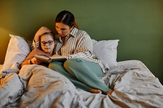 Minimal portrait of loving mother reading book to daughter with down syndrome at bedtime, copy space