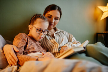 Portrait of little girl with down syndrome reading book at bedtime and relaxing with caring mother...
