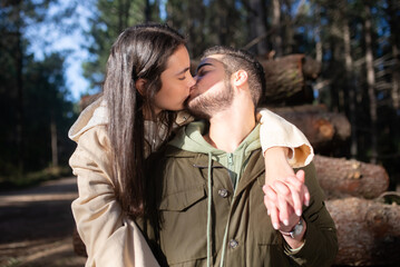 Fototapeta premium Portrait of young affectionate couple kissing in forest. Woman with dark hair and bearded man holding hands. Love, affection, relationship concept