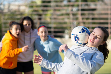 Fototapeta premium Happy school girls having fun with ball on stadium. Team of four joyous girlfriends preparing for football game training to win and demonstrating skills. Healthy lifestyle, leisure, team sport concept